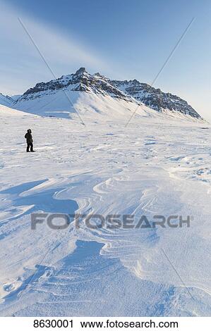 Stock Image - Man in a parka standing on the windswept snow, sastrugi patterns in the foreground, Soakpak Mountain in the distance, Anaktuvuk Pass, Gates of the Arctic National Park; Alaska, United States of America. Fotosearch