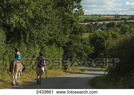 Stock Image - Mother with girl on donkey walking along a road; Normandy, France. Fotosearch