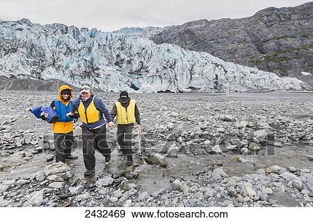 Stock Photo - Mother with two teenage sons wearing life jackets, crossing a stream in front of Shoup glacier and Chugach mountains, Shoup Bay State Marine Park, Prince William Sound; Valdez, Alaska, United States of America. Fotosearch