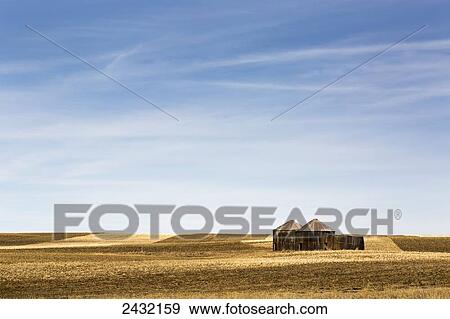 Stock Photo - Old wooden grain bins in a cut stubble field with blue sky and hazy clouds; Alberta, Canada. Fotosearch