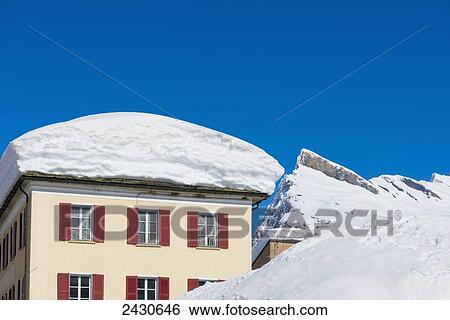 Stock Photograph - Pile of snow on a rooftop with snow covered mountains in the background and blue sky; San Bernardino, Grisons, Switzerland. Fotosearch