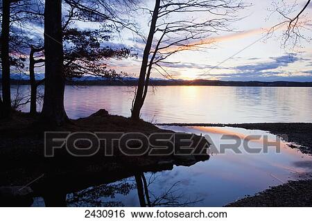 Stock Photograph - Reflection of sky and trees along the shoreline at sunset; Starnberger See, Bavaria, Germany. Fotosearch