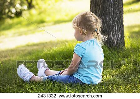 Stock Image - Toddler girl sitting in a park beside a tree; Edmonton, Alberta, Canada. Fotosearch