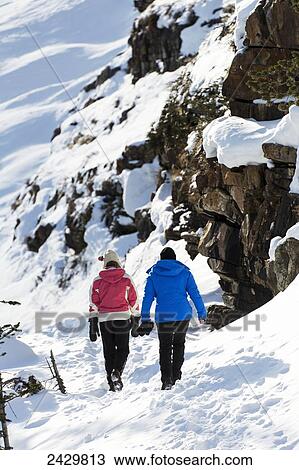 Stock Image - Two female hikers along a snow covered trail and snow covered rock cliff; Lake Louise, Alberta, Canada. Fotosearch