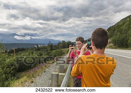 Stock Image - Two teenage boys on the side of the Glen highway taking photos of the Chugach mountains with cell phones; Alaska, United States of America. Fotosearch