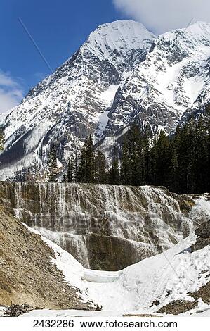 Waterfalls over rock cliff framed by snow covered rocks with snow covered mountain in the background and blue sky; Field, British Columbia, Canada View Large Photo Image Stock Photograph - Waterfalls over rock cliff framed by snow covered rocks with snow covered mountain in the background and blue sky; Field, British Columbia, Canada. Fotosearch