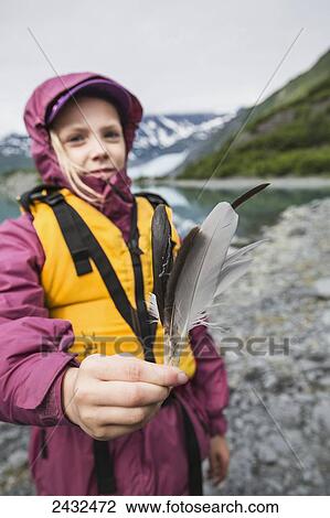 Stock Image - Young girl in yellow lifejacket displaying some bird feathers standing in front of Shoup glacier and Chugach mountains, Shoup Bay State Marine Park, Prince William Sound; Valdez, Alaska, United States of America. Fotosearch