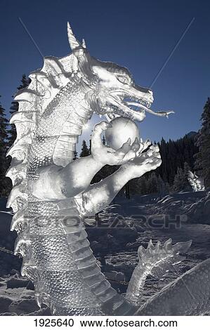 Stock Image - A Backlit Ice Sculpture Of A Dragon Holding A Sphere With Blue Sky And Trees In The Background; Lake Louise, Alberta, Canada. Fotosearch