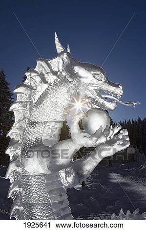 Stock Image - A Backlit Ice Sculpture Of A Dragon Holding A Sphere With Blue Sky And Trees In The Background And A Sunburst; Lake Louise, Alberta, Canada. Fotosearch
