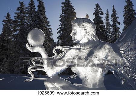 Stock Photo - A Backlit Ice Sculpture Of A Winged Fairy Holding A Sphere With Blue Sky And Trees In The Background; Lake Louise, Alberta, Canada. Fotosearch