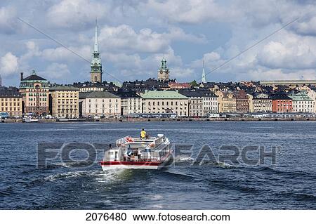 Stock Image - A Boat Traveling Through The Water And Buildings Along The Water's Edge; Stockholm Sweden. Fotosearch