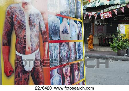 Stock Image - A Buddhist monk on his morning alms round passes a large advertisement for a tattoo studio; Bangkok, Thailand. Fotosearch