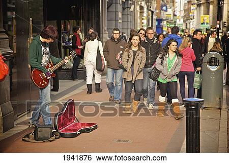 Stock Photograph - A Busker Playing A Guitar On The Sidewalk As Pedestrians Walk By On Grafton Street; Dublin Ireland. Fotosearch