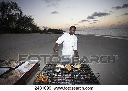 Stock Image - A chef cooks on a grill on the beach at sunset; Vamizi Island, Mozambique. Fotosearch