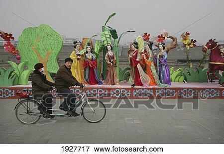 A Chinese Couple Riding A Tandem Bicycle Past A Traditional Display, Near The Old City Wall, Xian, Xi'an, Capital Of Shaanxi Province, China View Large Photo Image Stock Photo - A Chinese Couple Riding A Tandem Bicycle Past A Traditional Display, Near The Old City Wall, Xian, Xi'an, Capital Of Shaanxi Province, China. Fotosearch