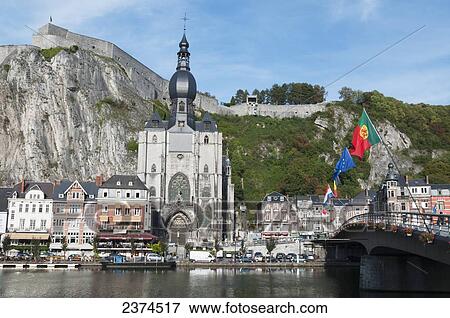 Stock Photo - A church and other buildings along the water and flags flying; Dinant, Belgium. Fotosearch