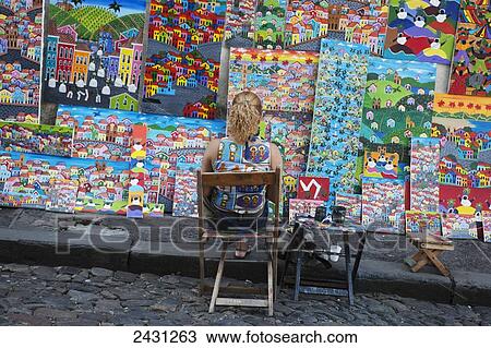 Stock Image - A cobbled shopping street in the historical centre of the old colonial city, with an artist working in front of a display of her art; Salvador, Brazil. Fotosearch