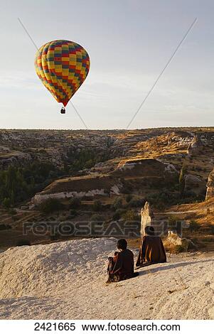 Stock Photography - A colourful hot air balloon in flight as two people sit on a rock to watch; Goreme, Cappadocia, Turkey. Fotosearch