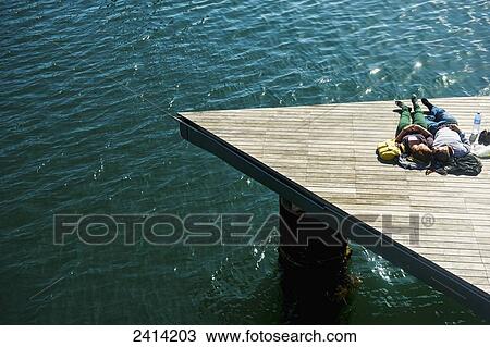 Stock Image - A couple laying on a wooden dock at the water's edge in the warm sunshine; Barcelona, Spain. Fotosearch
