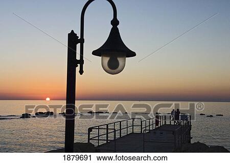 Stock Image - A Couple On A Small Pier Watch The Sun Setting At Sea. Fotosearch