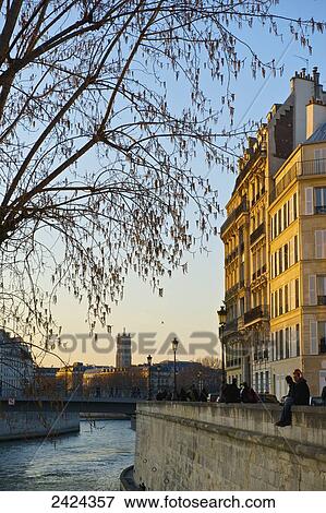 Stock Photo - A couple sit together on a canal wall with sunset sunlight cast on the buildings; Paris, France. Fotosearch