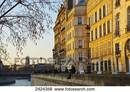 Stock Photo - A couple sit together on a canal wall with sunset sunlight cast on the buildings; Paris, France. Fotosearch