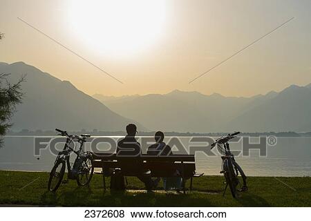 Stock Photo - A Couple Sits On A Park Bench At The Water's Edge With Their Bicycles At Sunset; Locarno, Ticino, Switzerland. Fotosearch