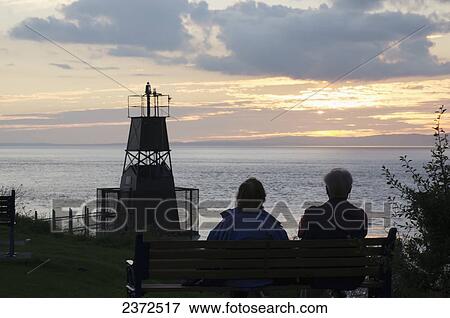 Stock Photo - A couple sits on the bench looking out over the water at sunset; Bristol, England. Fotosearch