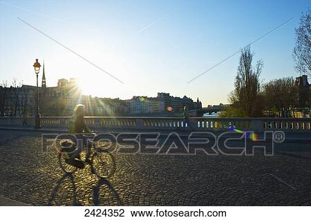 Stock Image - A cyclist rides on a street across a canal at sunrise; Paris, France. Fotosearch