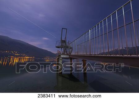 Stock Image - A diving board on the edge of tranquil Lake Maggiore at nighttime; Ascona, Ticino, Switzerland. Fotosearch