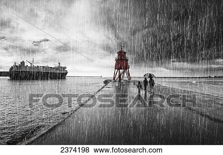 Stock Photo - A family walking in the pouring rain along harbour beach with Groyne Lighthouse and a ship; South Shields, Tyne and Wear, England. Fotosearch