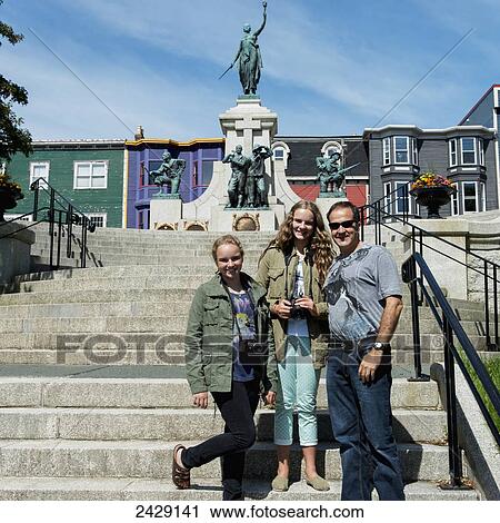 Stock Image - A father and his two daughters posing on steps in front of statues and colourful buildings; St. John's, Newfoundland, Canada. Fotosearch