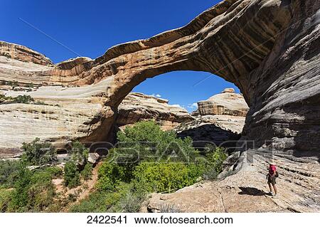 Stock Image - A female hiker looks at the rock bridge named Sipapu in the Natural Bridges National Monument; Utah, United States of America. Fotosearch