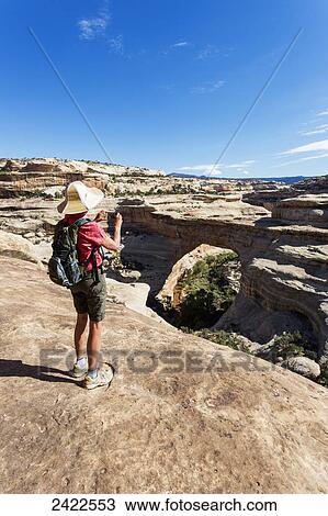 Stock Image - A female hiker takes a photo with a smart phone of the rock bridge named Sipapu in the Natural Bridges National Monument; Utah, United States of America. Fotosearch