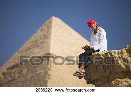Stock Image - A Female Tourist Reads A Guide Book While Sitting In Front Of The Pyramids Of Giza Near Cairo; Giza Egypt. Fotosearch