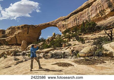 Stock Photograph - A female tourist takes a photo with a smart phone of the rock bridge named Owachomo in the Natural Bridges National Monument; Utah, United States of America. Fotosearch