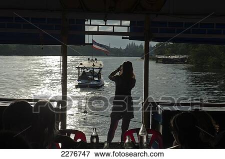 Stock Photo - A gap year student takes a photograph from a floating restaurant of Bridge over The River Kwai; Kanchanaburi, Thailand. Fotosearch
