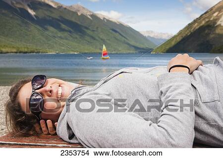Picture - A Girl Lies Down On A Picnic Table And Enjoys The Sun With A Sailboat In Lake Rotoiti In The Background, Tasman Region; New Zealand. Fotosearch