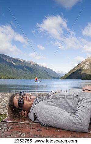 Stock Photography - A Girl Lies Down On A Picnic Table And Enjoys The Sun With A Sailboat In Lake Rotoiti In The Background, Tasman Region; New Zealand. Fotosearch