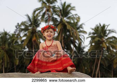 Stock Photography - A girl on a rock in a red dress with flowers in her hair, Patnum beach, Goa, India.. Fotosearch