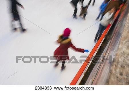 Stock Image - A girl wearing a plaid red and black coat with a pink hat skating at the seasonal skating rink in Rockafeller Centre; New York City, New York, United States of America. Fotosearch