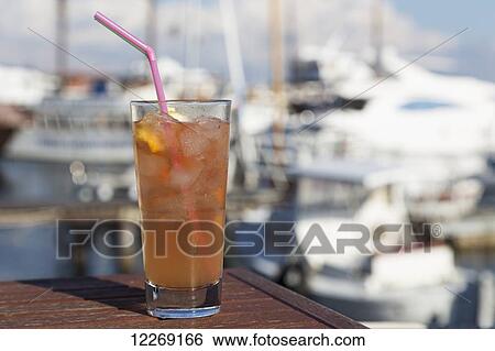 Stock Photograph - A glass of iced tea on a table with boats in the harbour in the background; Paphos, Cyprus. Fotosearch