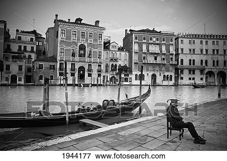 Stock Photo - A Gondolier Sits On A Chair On The Promenade Beside A Moored Gondola; Venice Venezia Italy. Fotosearch