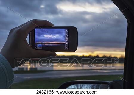 Stock Photograph - A hand holds a smart phone to show the image of Dorney Lake at sunset; Buckinghamshire, England. Fotosearch