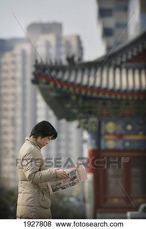 Stock Photo - A Local Woman Reads A Newspaper, In The Background There Is A Contrast Of Old And New, A Traditional Chinese Structure Beside Modern Skyscrapers, Beijing, China. Fotosearch