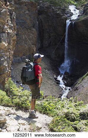 Stock Image - A male hiker overlooking a waterfall on a trail; Waterton, Alberta, Canada. Fotosearch