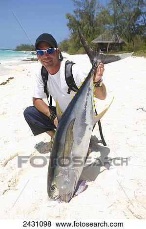 Stock Photo - A man crouches in the white sand holding a large fish; Vamizi Island, Mozambique. Fotosearch