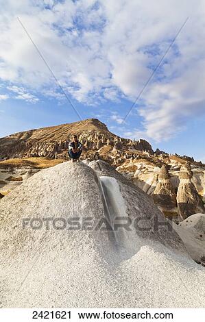 Stock Image - A man crouches on a large rock in a landscape of fairy chimneys; Cappadocia, Turkey. Fotosearch