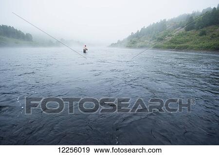 Stock Photo - A man fly fishing for brook trout on a wide misty wilderness river; Ontario, Canada. Fotosearch