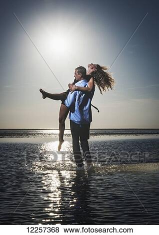 A man holds a woman in the shallow water along the coast looking out at the ocean; Tarifa, Cadiz, Andalusia, Spain View Large Photo Image Stock Photo - A man holds a woman in the shallow water along the coast looking out at the ocean; Tarifa, Cadiz, Andalusia, Spain. Fotosearch